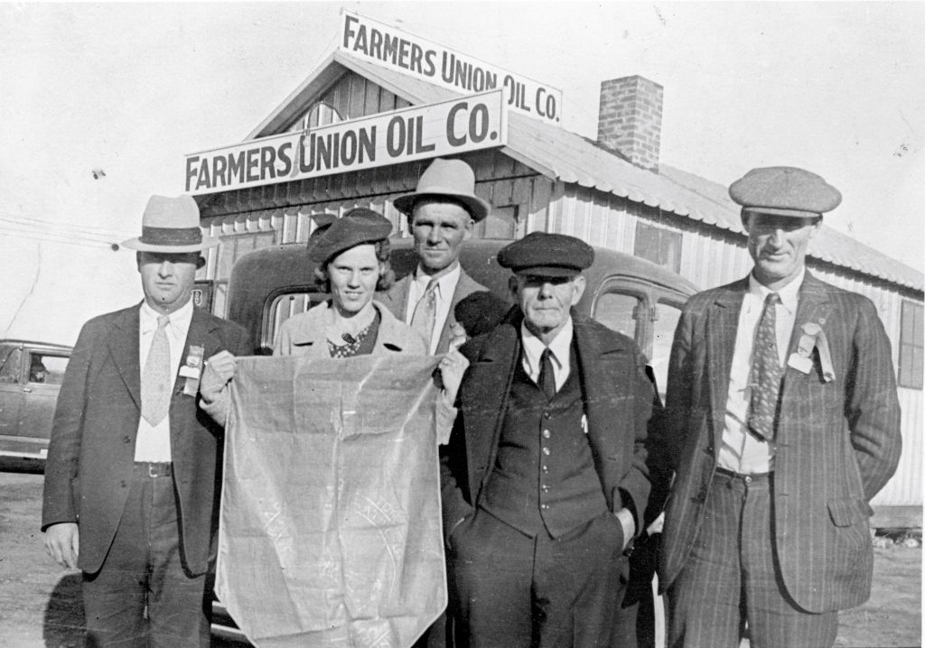 Five Farmers Union members standing in front of the Farmers Union Oil Co., with two members holding a Farmers Union banner.
